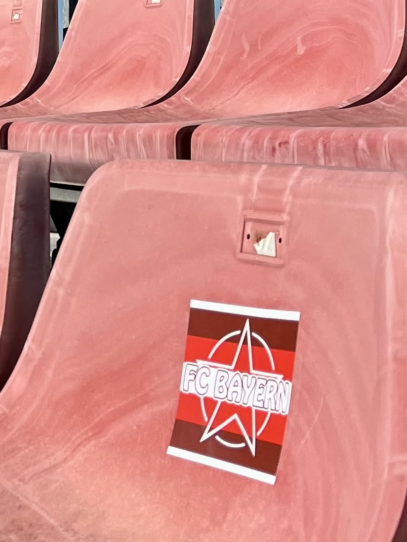 A rectangular sticker featuring a design with alternating horizontal stripes of red and dark brown/maroon. Overlaid on these stripes is a prominent white outline of a five-pointed star, partially encircled by a white ring. The text "FC BAYERN" is written in bold, white, sans-serif capital letters across the center of the sticker, overlapping the star and stripes. The sticker appears to be slightly peeling at its bottom edge, adhering to a dusty, worn pink stadium seat.