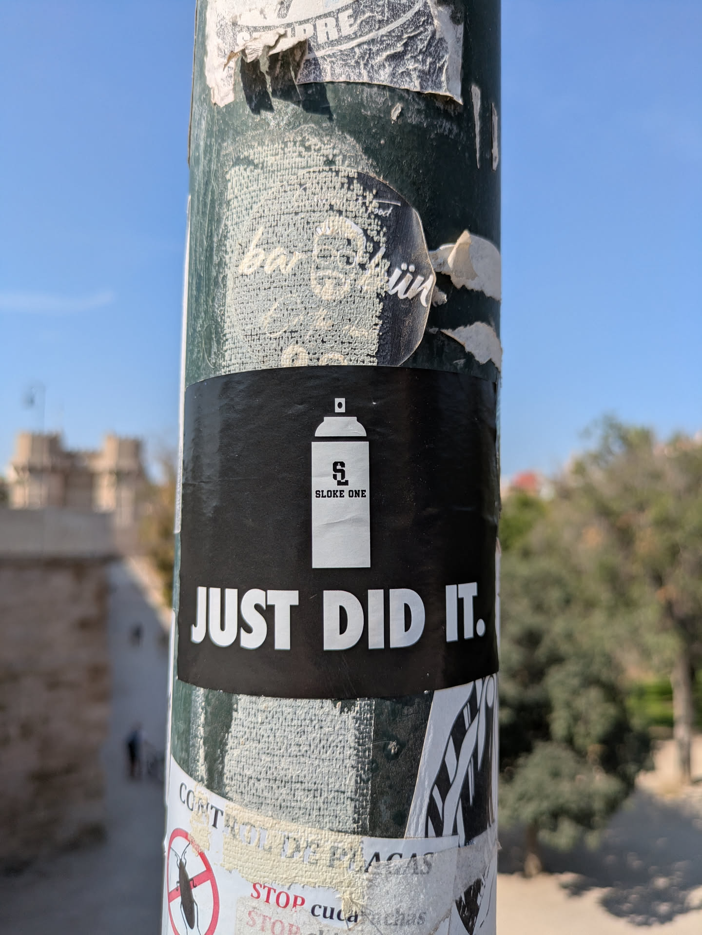 The sticker features a white spray can with the letters 'S' and '2' inside, followed by the text 'SLOKE ONE' and 'JUST DID IT.' The sticker is black with white text and is placed on a pole with other stickers.