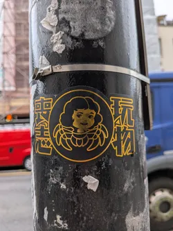 The sticker is predominantly black with all design elements rendered in a vibrant golden yellow. It features a central circular emblem containing a stylized, smiling child's face with large, curly hair or a halo-like structure around its head. The face is simple, with dots for eyes and a curved line for a mouth, all in golden yellow. Flanking this central emblem are two vertical columns of Chinese characters, also in golden yellow outline and fill. The entire sticker has a clean, graphic design aesthetic, contrasting with the textured, worn black surface of the pole it's affixed to.