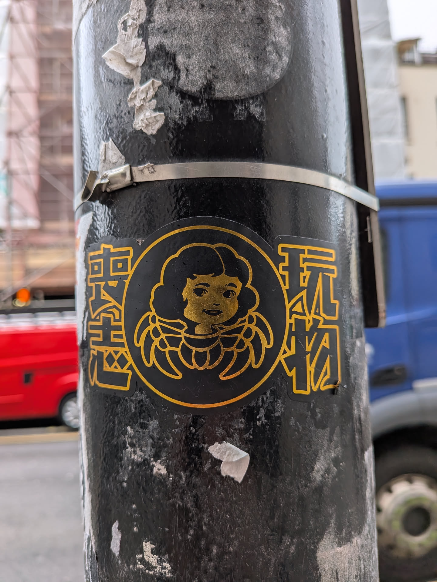 The sticker is predominantly black with all design elements rendered in a vibrant golden yellow. It features a central circular emblem containing a stylized, smiling child's face with large, curly hair or a halo-like structure around its head. The face is simple, with dots for eyes and a curved line for a mouth, all in golden yellow. Flanking this central emblem are two vertical columns of Chinese characters, also in golden yellow outline and fill. The entire sticker has a clean, graphic design aesthetic, contrasting with the textured, worn black surface of the pole it's affixed to.