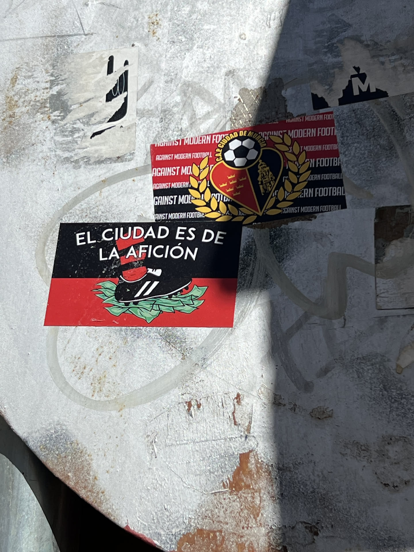 The primary sticker, centrally located, is rectangular with a horizontal split. The upper half is black, featuring white text that reads "EL CIUDAD ES DE LA AFICIÓN". The lower half is red and depicts a black football boot with white stripes, wearing a red and black striped sock, firmly planted on a pile of green leaves, which symbolize money. Behind and slightly above this sticker, a stack of smaller, identical red stickers is visible. These background stickers repeatedly display the white text "AGAINST MODERN FOOTBALL" and feature a central emblem: a football positioned above a heart-shaped shield (half red, half black) encircled by golden laurel wreaths. The shield also contains the text "CAP CIUDAD DE MURCIA" and various golden motifs. The overall aesthetic is bold and conveys a strong message related to football culture.