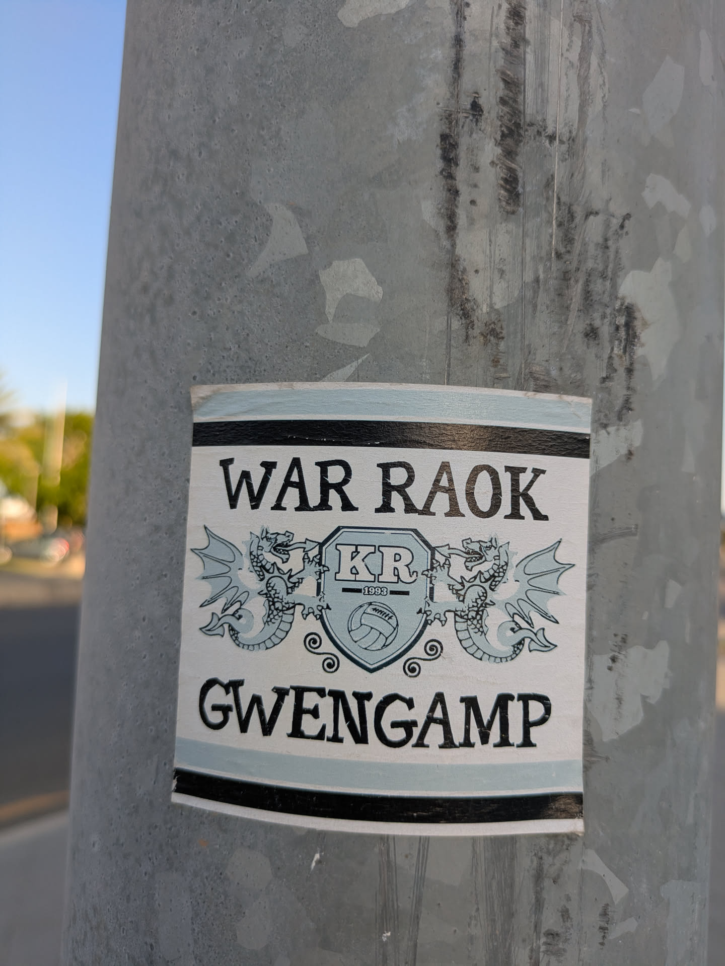 This is a rectangular sticker attached to a grey metal pole. The sticker features the words "WAR RAOK" at the top and "GWENGAMP" at the bottom, both in bold, black letters. In the center is an emblem with two dragons flanking a shield containing the letters "KR" above a volleyball. The year "1993" is printed beneath the letters.