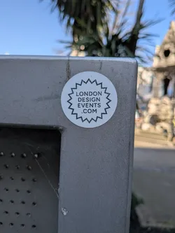 Street sticker The sticker is circular with a clean white background. It features a distinctive black, jagged, star-like border that frames the central text. Inside this border, the words "LONDON DESIGN EVENTS .COM" are stacked in three lines, rendered in a clear, sans-serif black font. The sticker is adhered to a textured, matte grey metal surface.