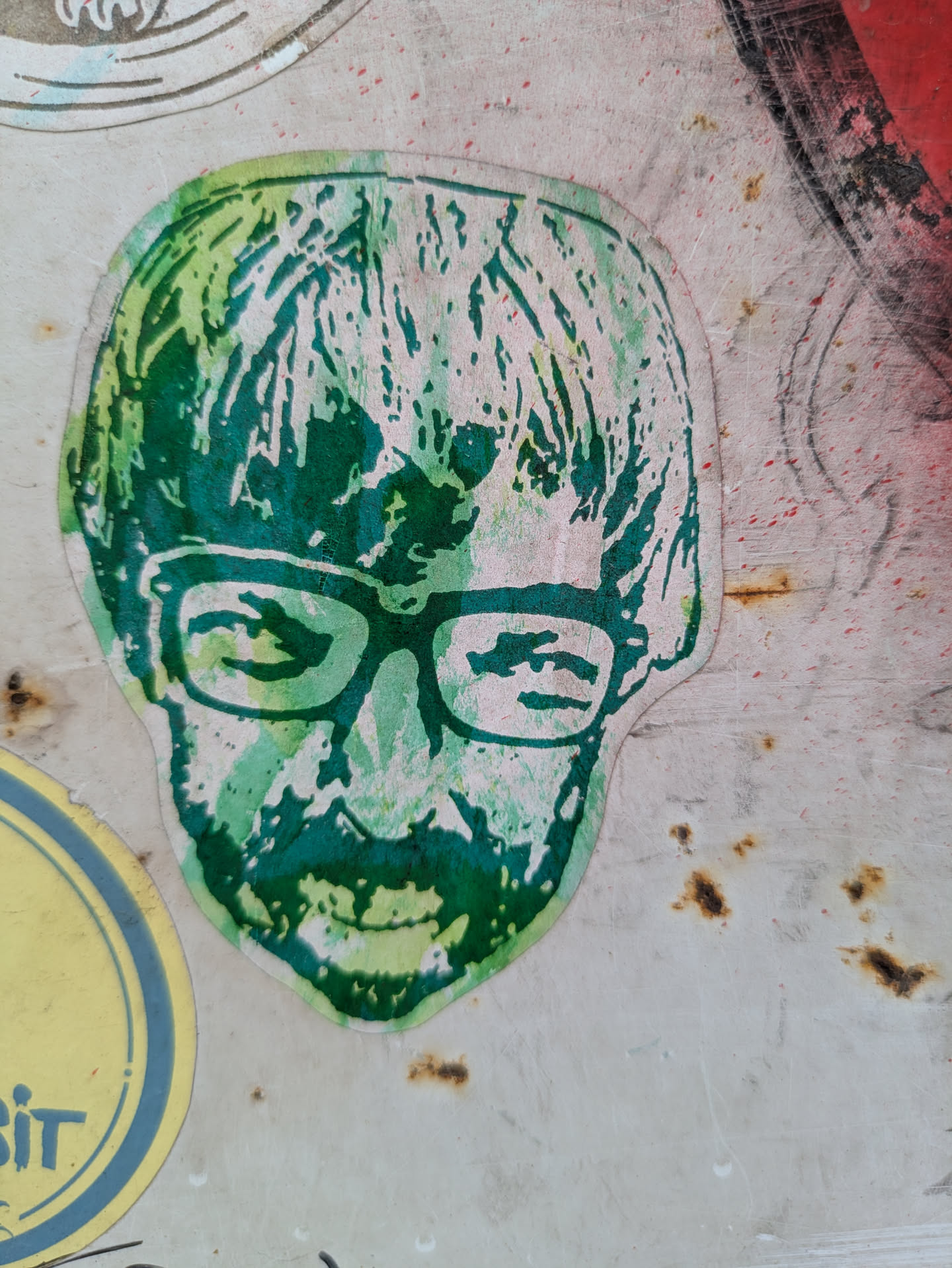 A green portrait sticker featuring the face of a man with glasses and a beard is adhered to a weathered surface. The sticker's colors are primarily shades of green, creating a vibrant contrast against the aged background. The portrait style is artistic, and the man's expression is neutral. The glasses add a distinct feature to his appearance.