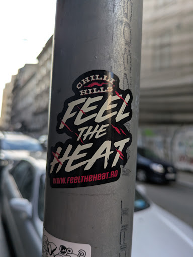 A black and white sticker adhered to a metal pole with the text "Chilli Hills Feel The Heat" and the website "www.feeltheheat.ro". The text is stylized with red accents.