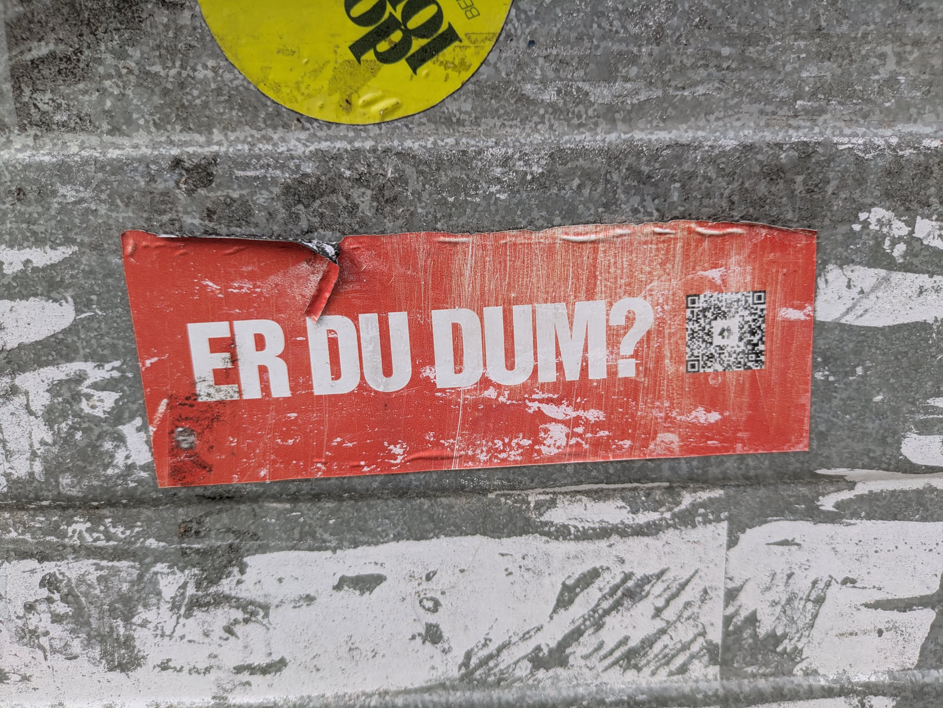 A rectangular, red sticker affixed to a rough, grey surface. The sticker is worn and creased, with some white scuff marks. It features bold white capital letters that read "ER DU DUM?" (Danish for "ARE YOU STUPID?") on the left side, and a black and white QR code on the right.