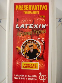 Street sticker A rectangular sticker, predominantly red, featuring a background pattern of strawberries. At the top, 'PRESERVATIVO' is in large yellow letters, followed by 'TRANSPARENTE'. Below this, 'LATEXIN®' is in white, and 'Aroma Fresa' in gold/brown. The central element is a circular emblem with an orange-yellow background and faint skull patterns, depicting a bald man with a long grey beard and sunglasses, wearing a black t-shirt and holding a beer bottle. The text around the circle reads 'CERTIFICADO POLLA VIEJA'. To the right of the emblem, 'CE' and '0318' are visible. A yellow rectangle below the emblem contains 'VENTA EN FARMACIAS' in red. The bottom section features 'GARANTIA DE CALIDAD, SEGURIDAD Y EFICACIA' in white, accompanied by a white symbol of intertwined male and female gender signs.