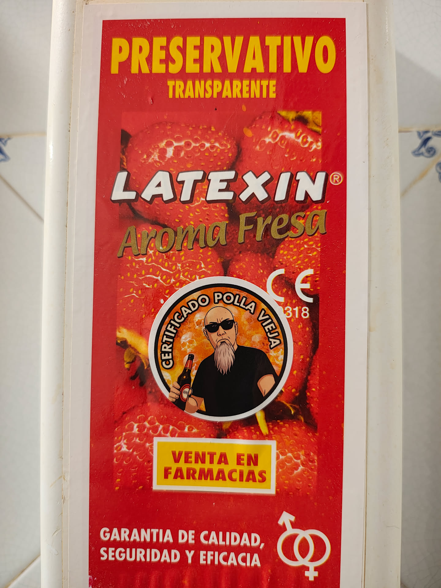 A rectangular sticker, predominantly red, featuring a background pattern of strawberries. At the top, 'PRESERVATIVO' is in large yellow letters, followed by 'TRANSPARENTE'. Below this, 'LATEXIN®' is in white, and 'Aroma Fresa' in gold/brown. The central element is a circular emblem with an orange-yellow background and faint skull patterns, depicting a bald man with a long grey beard and sunglasses, wearing a black t-shirt and holding a beer bottle. The text around the circle reads 'CERTIFICADO POLLA VIEJA'. To the right of the emblem, 'CE' and '0318' are visible. A yellow rectangle below the emblem contains 'VENTA EN FARMACIAS' in red. The bottom section features 'GARANTIA DE CALIDAD, SEGURIDAD Y EFICACIA' in white, accompanied by a white symbol of intertwined male and female gender signs.