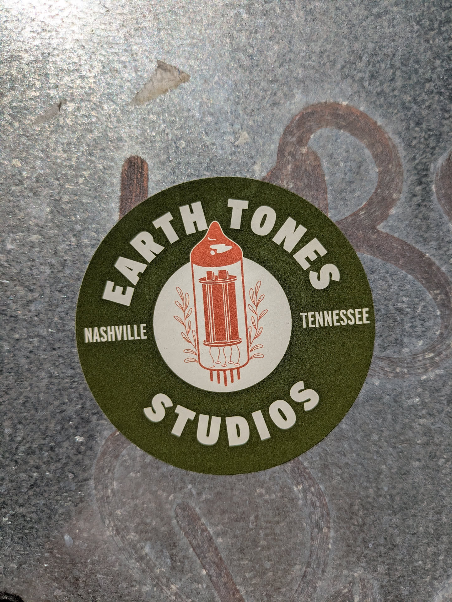 A circular sticker with a dark olive green background. The center features a white circle enclosing an illustration of an orange-red vintage vacuum tube, flanked by two small leafy sprigs. White bold text around the circle reads "EARTH TONES" at the top, "STUDIOS" at the bottom, "NASHVILLE" on the left, and "TENNESSEE" on the right. The sticker is affixed to a textured, possibly metallic or concrete surface.
