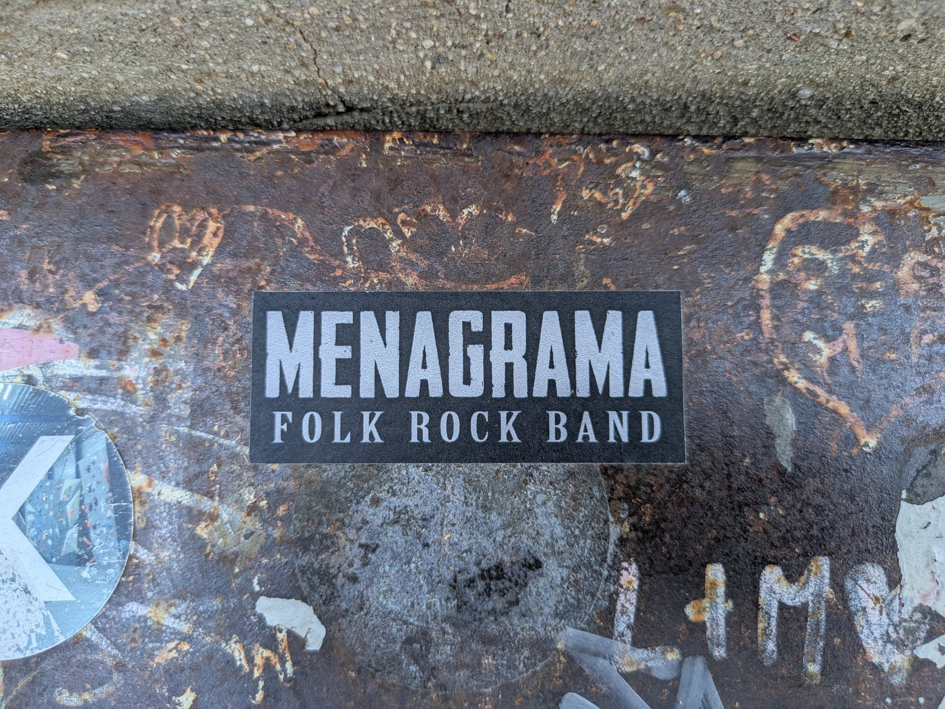 A black rectangular sticker adhered to a rusty, textured surface. The sticker features the band name "MENAGRAMA" in bold, white sans-serif letters, with the descriptor "FOLK ROCK BAND" in a smaller font beneath it. The surface shows signs of wear and tear, with streaks of rust and other markings adding to its aged appearance. The sticker is centrally positioned and contrasts against the corroded background.
