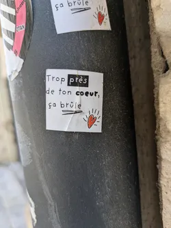 Street sticker A small, square sticker on a pole reads "Trop près de ton coeur, ça brûle" (Too close to your heart, it burns). A small red heart is drawn near the bottom right corner of the sticker.