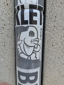 Street sticker This sticker depicts a cute, rounded fish character with cartoonish features. The fish is drawn in black and white, and to the right of the fish is the vertical Japanese text "魚の侵入" (sakana no shinnyū) which translates to "fish intrusion". The sticker is slightly damaged, with some parts torn off at the edges.