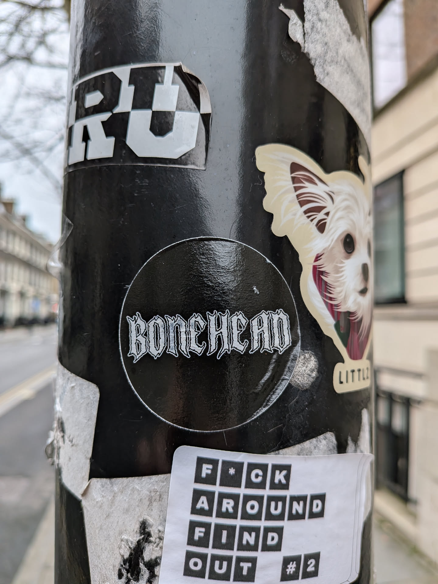 A circular black sticker prominently featuring the word 'BONEHEAD' in a stylized white gothic font. The letters are outlined and have a distressed, sharp-edged appearance, evoking a heavy metal or punk rock aesthetic. The sticker has a slightly glossy finish, reflecting ambient light, and is affixed to a dark, textured pole.