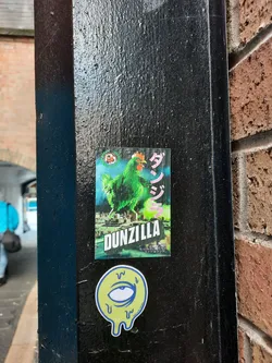 Street sticker The central sticker features a vibrant green chicken, depicted in a kaiju-like pose, standing over a stylized cityscape. The chicken has a red comb and wattle, and its wings are spread. Behind it, the city appears to be under a dramatic, possibly stormy, sky with hints of pink and blue. To the right of the chicken, large Japanese characters are visible in pink. Below the chicken, the word 'DUNZILLA' is prominently displayed in bold, white, block letters with a green outline. Smaller Japanese text is present at the bottom of this sticker. Below this main sticker is a smaller, circular sticker with a yellow background and a blue outline. It features a stylized eye with a blue iris and pupil, surrounded by dripping lines, giving it a melting appearance. The eye sticker also has some faint blue text around its bottom edge.