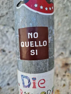 A brown square sticker with the text "NO QUELLO SI" on it,  attached to a metal pole. The text is in Italian and translates to "No That Yes".