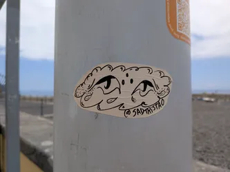 Street sticker An irregular beige sticker featuring a black ink drawing of a stylized face with large, sad eyes, enclosed in a wavy, cloud-like border. The handle '@SADTRISTAO' is written along the bottom right edge.