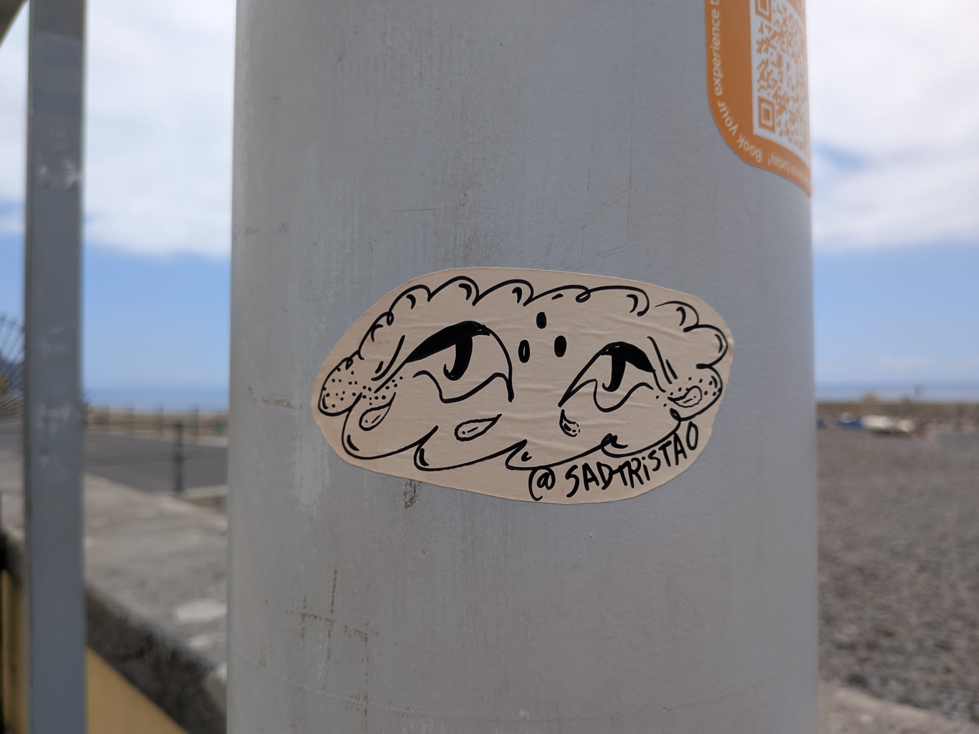 An irregular beige sticker featuring a black ink drawing of a stylized face with large, sad eyes, enclosed in a wavy, cloud-like border. The handle '@SADTRISTAO' is written along the bottom right edge.