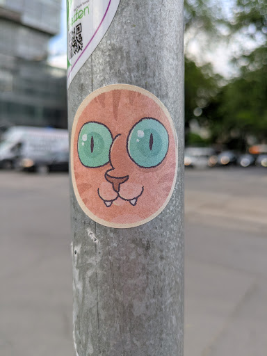 A round sticker on a metal pole features a cartoon cat face. The cat has orange fur, large turquoise eyes, a small nose, and a whimsical smile with two tiny teeth showing. The sticker has a light border and is adhered to the gray metal surface of the pole.