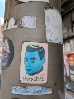 Street sticker A blue-toned sticker of a man's face is affixed to a gray pole. The sticker, which is slightly worn, features a man with sharp features and yellow eyes. Below the portrait, the text "jangojim" is written in a stylized font, possibly in Japanese.