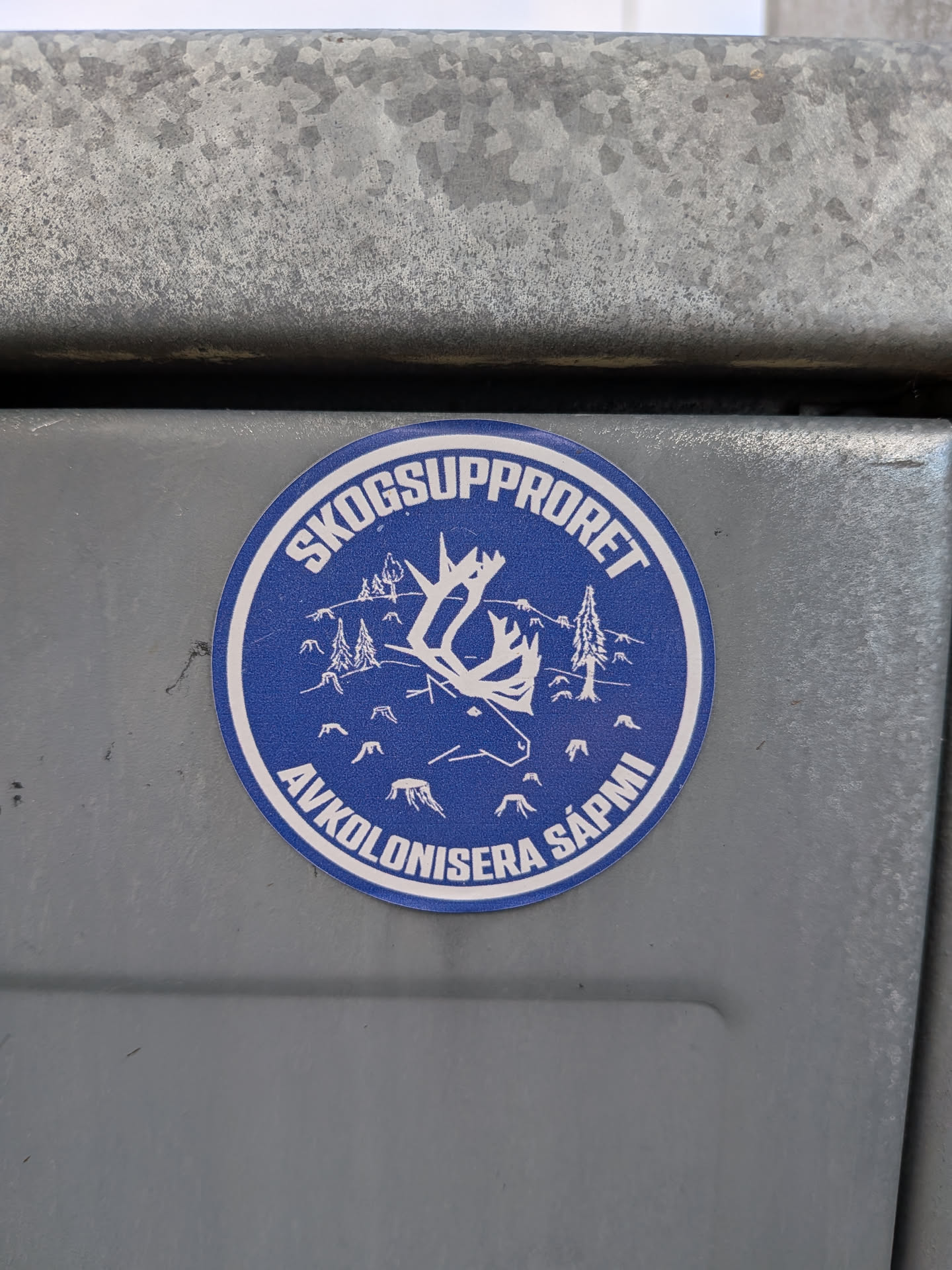 The sticker is circular with a blue background and white graphics. It features a stylized white outline of a reindeer's head and antlers in the center. Surrounding the reindeer are small white outlines of pine trees and tree stumps, suggesting a deforested or threatened landscape. The top arc of the circle contains the text 'SKOGSUPPRORET' in white capital letters, and the bottom arc contains 'AVKOLONISERA SÁPMI' in white capital letters. The entire design is enclosed by a thin white border.