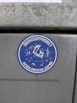 Street sticker The sticker is circular with a blue background and white graphics. It features a stylized white outline of a reindeer's head and antlers in the center. Surrounding the reindeer are small white outlines of pine trees and tree stumps, suggesting a deforested or threatened landscape. The top arc of the circle contains the text 'SKOGSUPPRORET' in white capital letters, and the bottom arc contains 'AVKOLONISERA SÁPMI' in white capital letters. The entire design is enclosed by a thin white border.