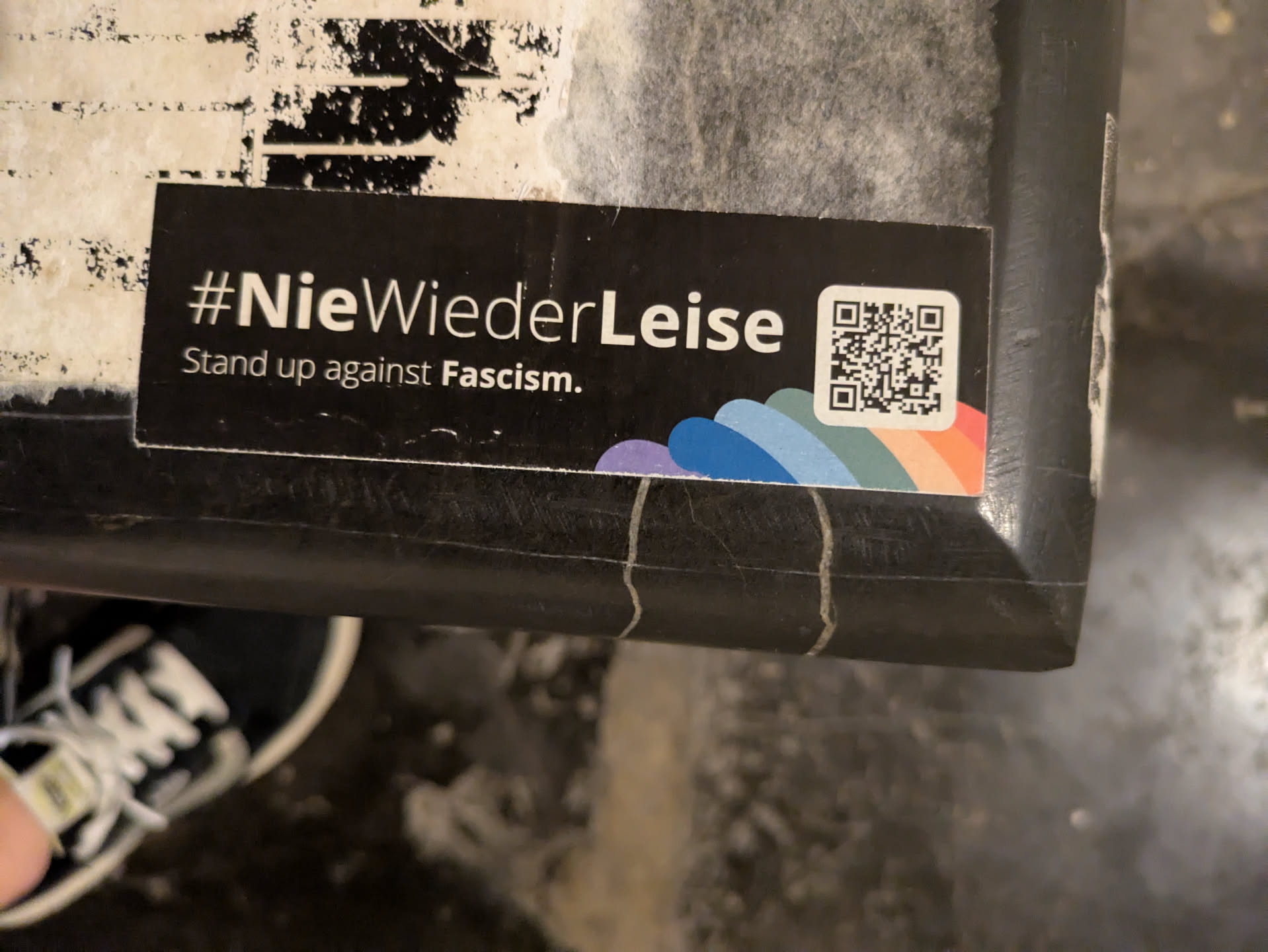 Street sticker The sticker is black with white text and a small rainbow graphic. A QR code is located on the right side of the sticker.
