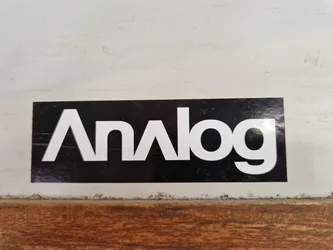Street sticker A black and white sticker with the word "Analog" written in a stylized font. The sticker is affixed to a light-colored surface that looks like painted wood or metal. The background is slightly dirty or worn.