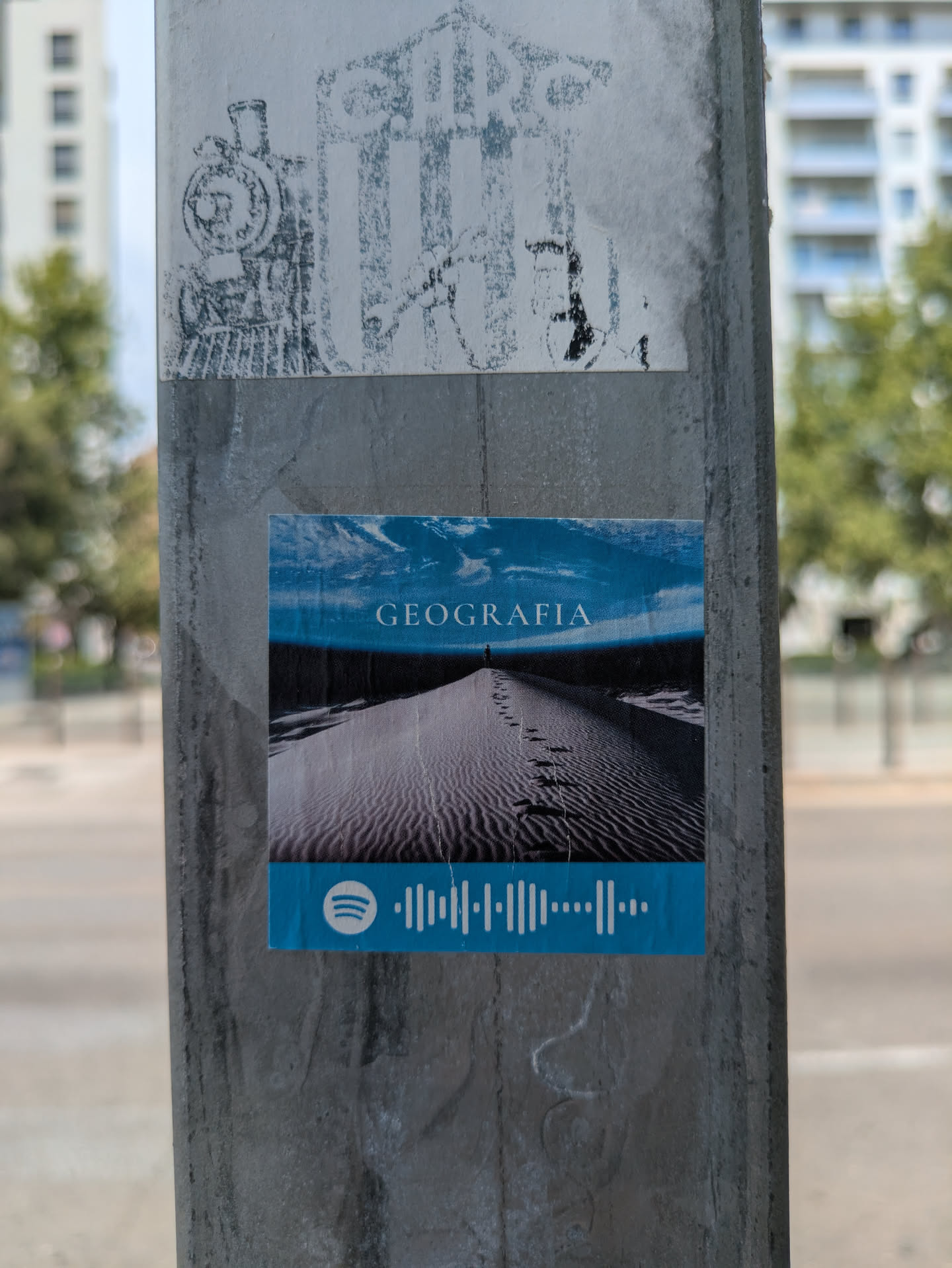 The sticker features an image of sand dunes under a blue sky, with a person standing in the distance. The word "GEOGRAFIA" is printed at the top of the image and a Spotify code at the bottom.