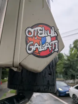 Street sticker A round sticker with the text 'OȚELUL GALAȚI' in dripping white graffiti font. The background is split, showing an orange-red pattern on the left and red, white, and blue segments on the right.