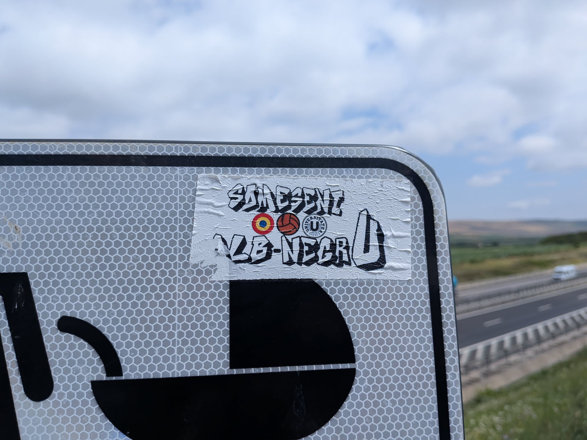 A rectangular white sticker on a road sign. It features black graffiti-style text reading 'SOMESENI' and 'ALB-NEGRU U'. Three central icons depict a Romanian tricolor target, a vintage football, and the Universitatea Cluj club crest.