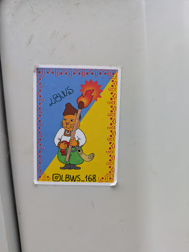 A sticker features a cat dressed as a cossack holding a torch in front of a blue and yellow background that mimics the Ukrainian flag. The frame of the image is decorated with Ukrainian folk art. The handle of the torch resembles a broom handle. The Instagram handle @LBWS_168 is on the bottom of the image.