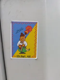 Street sticker A sticker features a cat dressed as a cossack holding a torch in front of a blue and yellow background that mimics the Ukrainian flag. The frame of the image is decorated with Ukrainian folk art. The handle of the torch resembles a broom handle. The Instagram handle @LBWS_168 is on the bottom of the image.