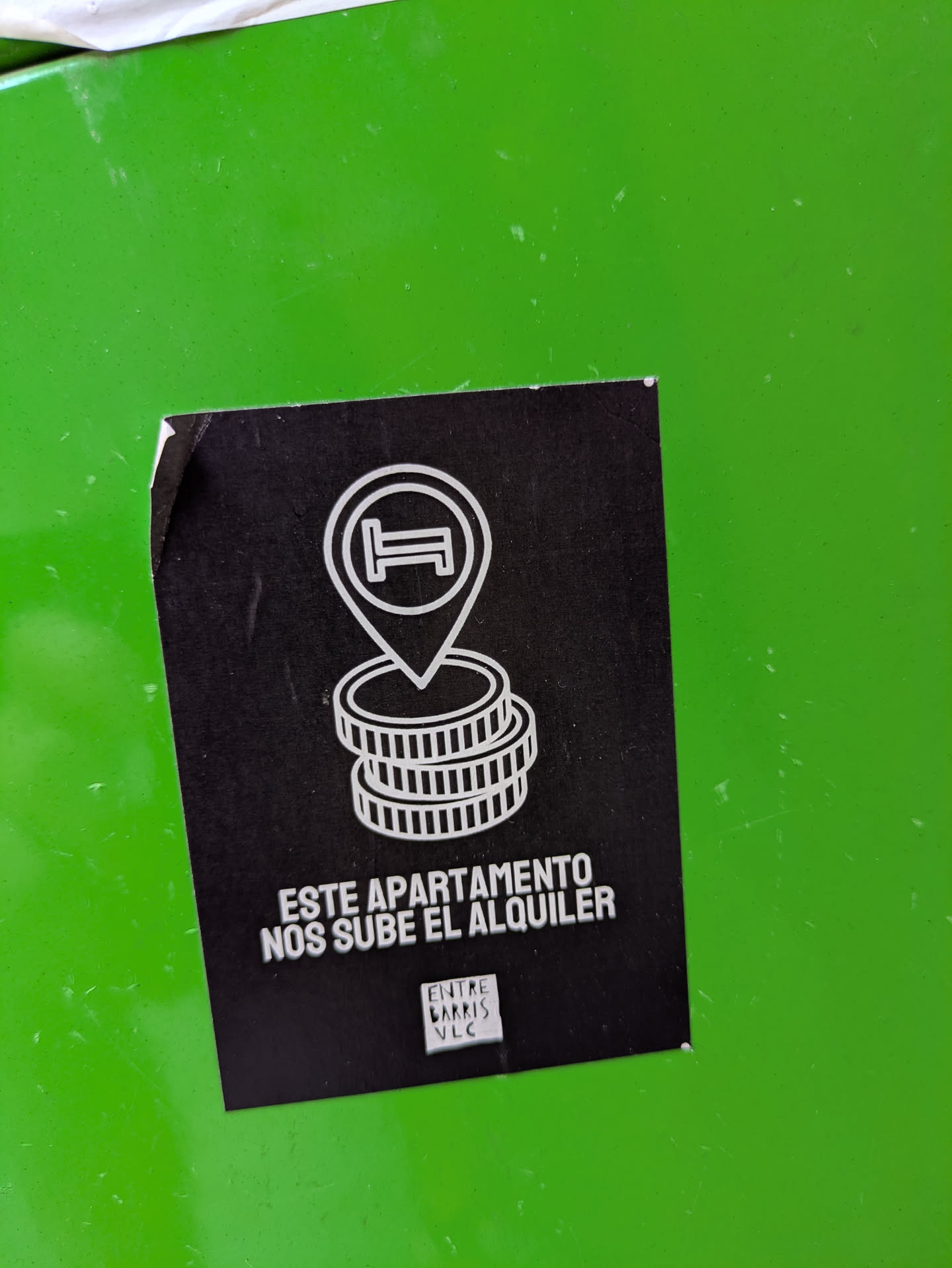 This black square sticker features a white outline illustration of a bed inside a map marker, positioned above stacks of coins, which is suggestive of increasing costs. The sticker includes the text "ESTE APARTAMENTO NOS SUBE EL ALQUILER", which translates to "THIS APARTMENT IS RAISING OUR RENT." The sticker also has the text "ENTRE BARRIS VLC"