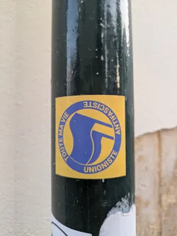 A yellow sticker with a blue logo featuring two flags inside a circle is attached to a dark-colored pole. The text "TOUTE MA VIE" and "ANTIFASCISTE UNIONISTE" surrounds the logo.