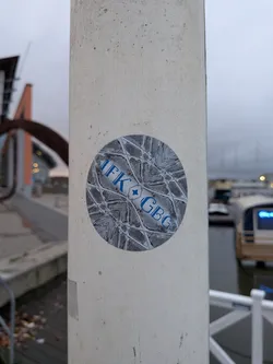 Street sticker The sticker is circular and features a complex, symmetrical geometric pattern in shades of gray and white. Superimposed on this pattern, in bold blue lettering, are the letters 'IFK' on the left and 'GBG' on the right, separated by a small blue diamond shape. The overall design is abstract and stylized, with the text clearly legible against the intricate background.