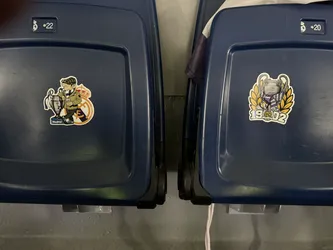 Street sticker The sticker features a cartoon illustration of a man, depicted as a football supporter, wearing a green jacket, a brown flat cap, and a purple and white striped scarf. He is leaning casually against a silver Champions League trophy, holding a small brown cup or drink in his right hand. Behind him, partially visible, is the classic Real Madrid club crest with its crown and intertwined 'M' and 'C'. Below the trophy, there's a small blue rectangular sign with 'Madrid' written in white. The sticker has a distinct white outline, suggesting a die-cut design, and is applied to a dark blue stadium seat.