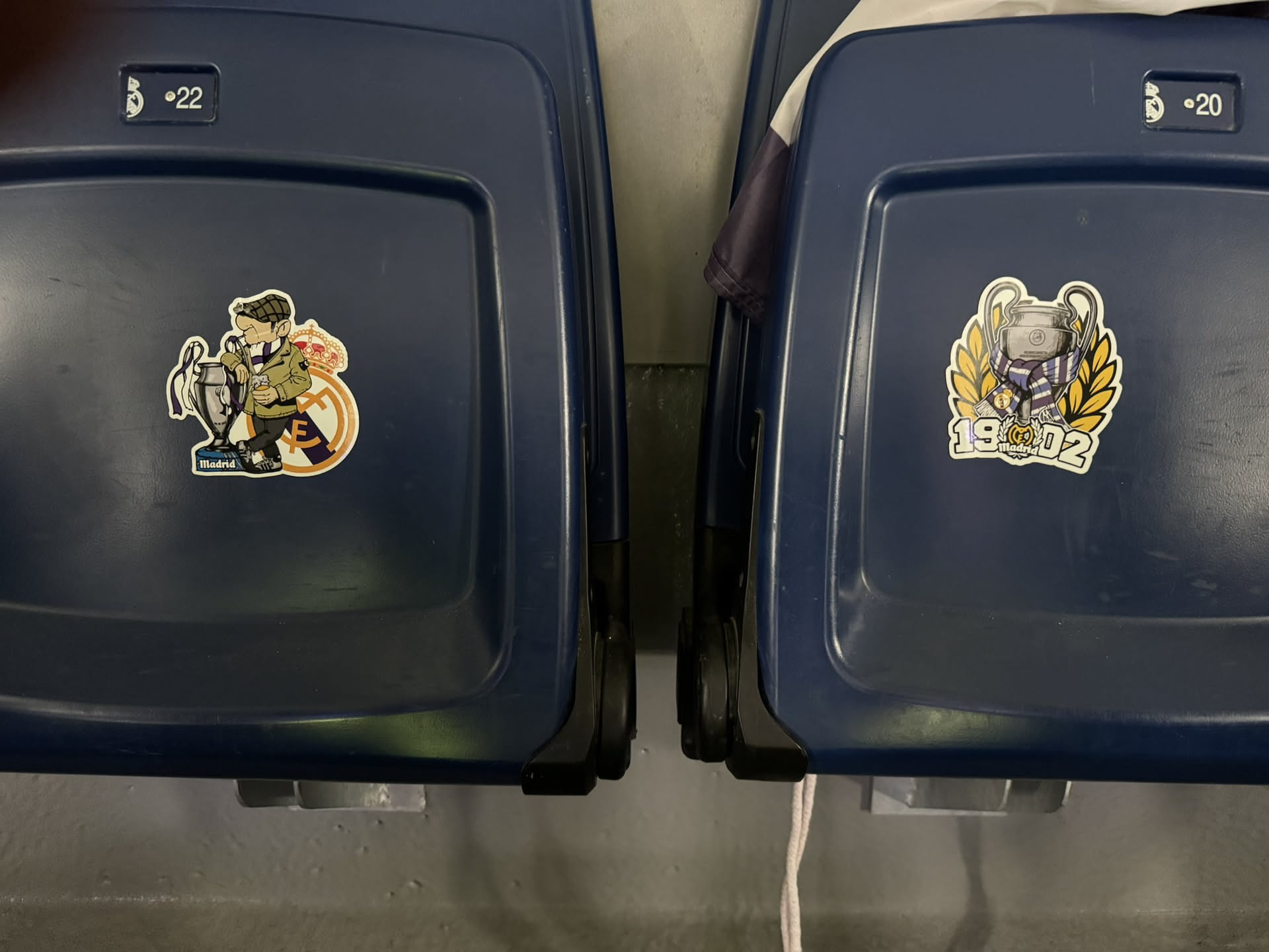 The sticker features a cartoon illustration of a man, depicted as a football supporter, wearing a green jacket, a brown flat cap, and a purple and white striped scarf. He is leaning casually against a silver Champions League trophy, holding a small brown cup or drink in his right hand. Behind him, partially visible, is the classic Real Madrid club crest with its crown and intertwined 'M' and 'C'. Below the trophy, there's a small blue rectangular sign with 'Madrid' written in white. The sticker has a distinct white outline, suggesting a die-cut design, and is applied to a dark blue stadium seat.