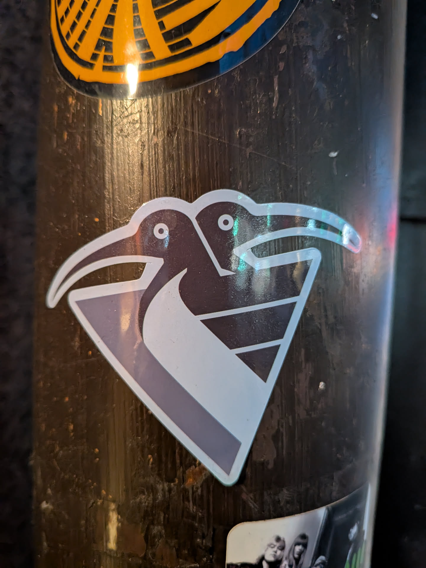 The sticker features two stylized kiwi birds facing left, with their heads overlapping. The birds are depicted in dark gray and white, with small black dots for eyes. They are positioned within a light purple and white geometric shape resembling a shield or a stylized diamond. The sticker has a holographic finish, causing iridescent reflections, particularly on the beaks and outlines of the birds. The background of the sticker is white, with a thin dark gray border.