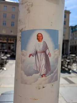 Street sticker The sticker features a smiling man with light-colored hair, depicted in a style reminiscent of classical religious art. He is dressed in a flowing white robe with a rope belt, standing barefoot on fluffy white clouds. A bright light emanates from behind him, creating a halo effect against a light blue sky background. The image is a clear parody or humorous take on traditional depictions of divine figures like Jesus Christ, by superimposing a contemporary, anachronistic face onto the iconic imagery. The sticker is placed on a weathered, light-colored pole in an outdoor urban setting.