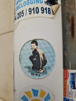 Street sticker A round sticker featuring a stylized illustration of a child squatting. The artwork is in a muted color palette, with the child depicted in dark clothing against a light blue background. The word "TONA" is printed below the illustration. The sticker shows some wear, indicating it has been affixed to a surface for a period of time.