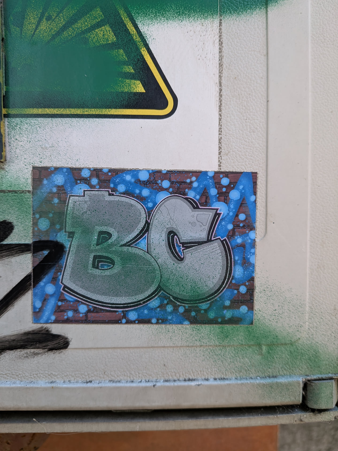 A rectangular sticker showing the letters 'BC' in a green-grey gradient bubble style. The background is a brick wall with blue spray paint bubbles.