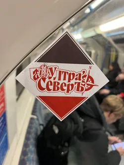 Street sticker A diamond-shaped sticker featuring a central white band with a distressed texture, bordered by a dark brown/black triangle at the top and a red triangle at the bottom, all outlined in white. The main text, 'Ультра Северъ' (Ultra Sever), is prominently displayed in bold red Cyrillic font across the white band. To the left of the text is an oval crest depicting a lion rampant holding a sword, with 'ЦСКА' (CSKA) written below it. To the right, a red compass rose points north, marked with an 'N'. The overall design is graphic and striking, using a primary color palette of red, white, and dark brown/black.