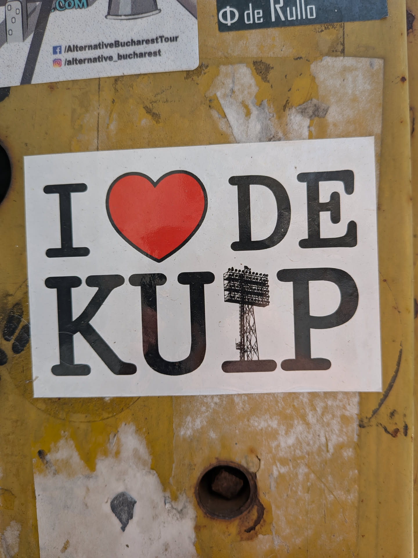 White rectangular sticker featuring the text 'I ❤️ DE KUIP' in black serif font. The heart is red with a black border, and the letter 'I' in 'KUIP' is replaced by a black silhouette of a stadium floodlight tower.