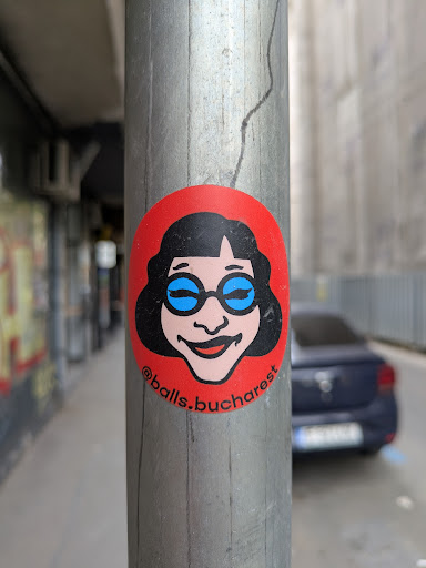 A round sticker on a metal pole shows a cartoon woman with black hair, blue glasses, and a smile. The sticker has a red background and the text '@balls.bucharest' is printed on it.
