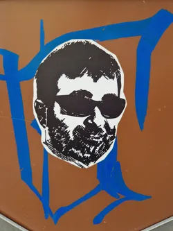 A black and white sticker depicting a man wearing sunglasses. The style is reminiscent of stencil art or a graphic design print. It is affixed to a brown surface with blue painted markings around it. 