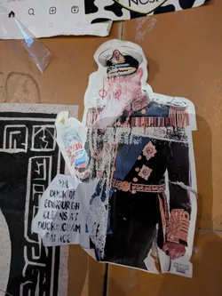 Street sticker The sticker features an image of the Duke of Edinburgh in full royal regalia, but with the humorous addition of holding a bottle of "TTO on Total, Cão Total" cleaning product. This implies he's cleaning Buckingham Palace, a playful take on his royal duties. The sticker also includes textual elements to reinforce the joke.