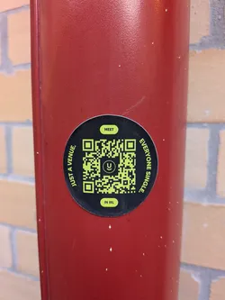 A circular black sticker with a bright green QR code at its center. The QR code has a small, stylized 'U' logo within it. Text is arranged around the QR code in a circular fashion. At the top, in a small green oval, is the word 'MEET'. Along the left side of the circle, in green text, reads 'JUST A VENUE.'. Along the right side, also in green text, reads 'EVERYONE SINGLE.'. At the bottom, in another small green oval, is the text 'IN IRL.'. The sticker is affixed to a textured red surface.