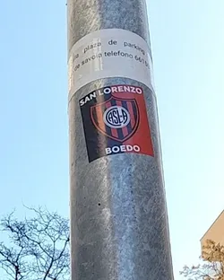 Street sticker A rectangular sticker featuring the crest of Club Atlético San Lorenzo de Almagro. The crest is a shield shape, diagonally divided into a black half on the left and a red half on the right. In the center, a circular emblem with the intertwined white letters 'CASLA' (Club Atlético San Lorenzo de Almagro) on a blue background is visible, encircled by a white ring. Above the crest, in an arc, the text 'SAN LORENZO' is written in white capital letters. Below the crest, the word 'BOEDO' is displayed in white capital letters. The sticker is affixed to a grey metal pole.