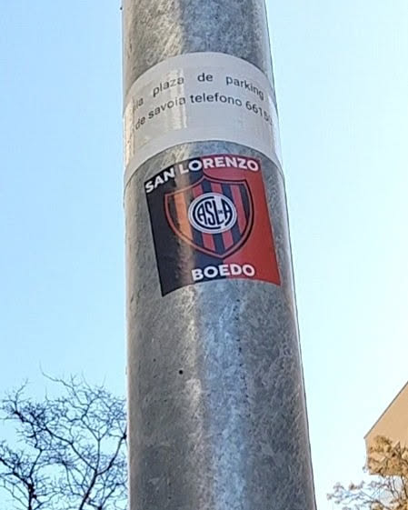 A rectangular sticker featuring the crest of Club Atlético San Lorenzo de Almagro. The crest is a shield shape, diagonally divided into a black half on the left and a red half on the right. In the center, a circular emblem with the intertwined white letters 'CASLA' (Club Atlético San Lorenzo de Almagro) on a blue background is visible, encircled by a white ring. Above the crest, in an arc, the text 'SAN LORENZO' is written in white capital letters. Below the crest, the word 'BOEDO' is displayed in white capital letters. The sticker is affixed to a grey metal pole.
