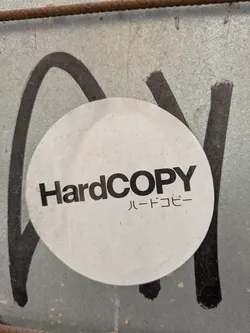 Street sticker The image shows a round, white sticker with the text 'HardCOPY' in bold, black letters. Below that, there is Japanese text that also translates to 'Hard Copy'. The sticker is attached to a corrugated metal surface which is also covered with black graffiti.